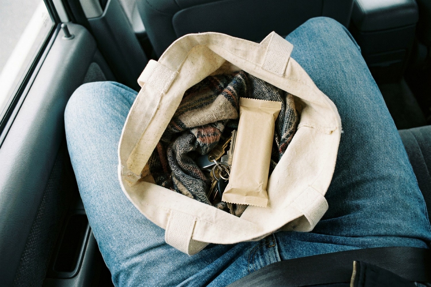 An open canvas tote bag resting on a lap in the passenger seat of a parked car, holding a folded plaid scarf, a set of keys and a plain paper-wrapped bar
